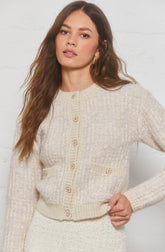 A woman wearing a cream-colored, textured knit cardigan with a subtle checkered pattern. The cardigan features gold decorative buttons along the front and on two small pockets at the waist. The cardigan has a round neckline and ribbed cuffs at the sleeves and hem.